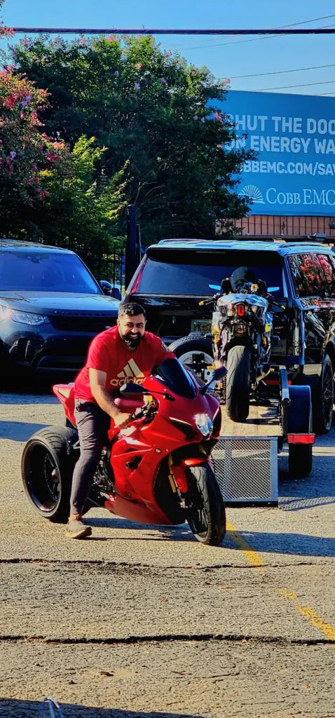 Man in a red shirt straddling a red Ducati-style superbike in a parking lot, with a black SUV and loaded motorcycle trailer behind him.