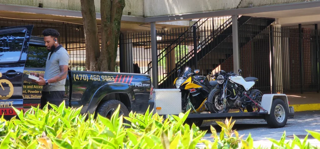 A man stands with a clipboard beside the branded Diaz Tacoma and open trailer loaded with a yellow sportbike and a white cafe racer, under a parking deck.