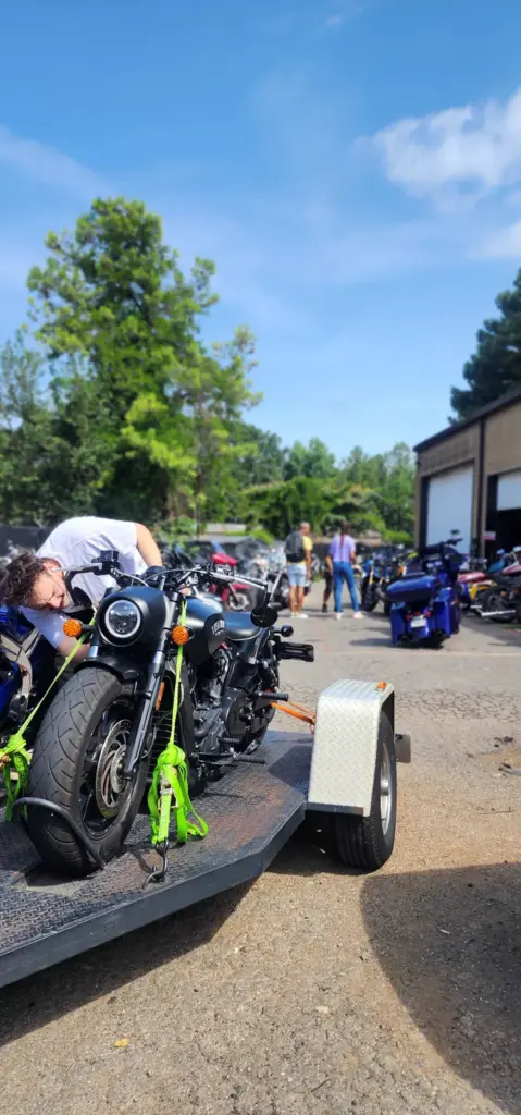A black motorcycle is strapped on a trailer in a sunny lot, surrounded by people and other motorcycles.