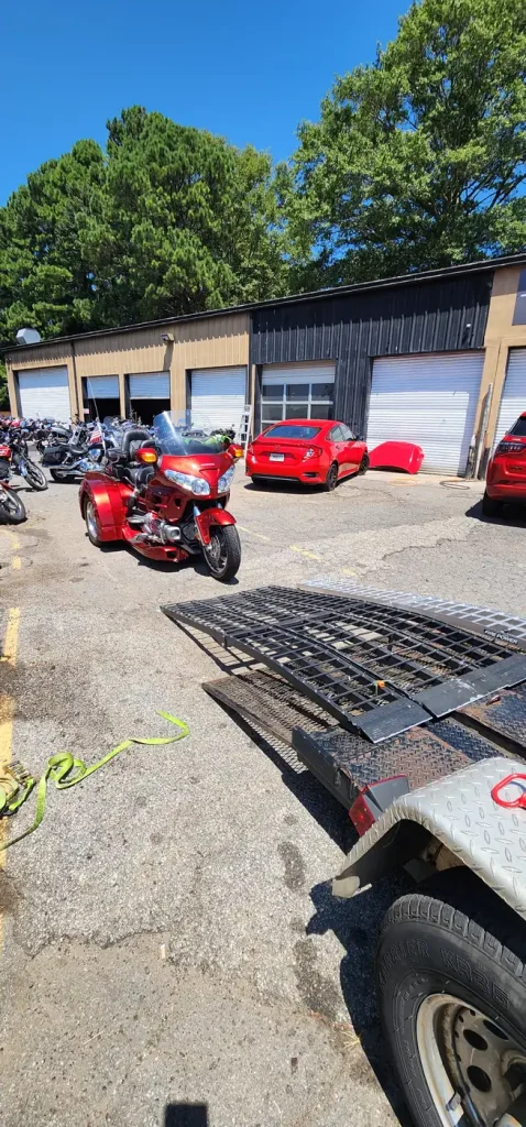 Open trailer with folding ramp and yellow ratchet straps on the ground, red Honda Gold Wing trike and rows of bikes outside the shop bays.