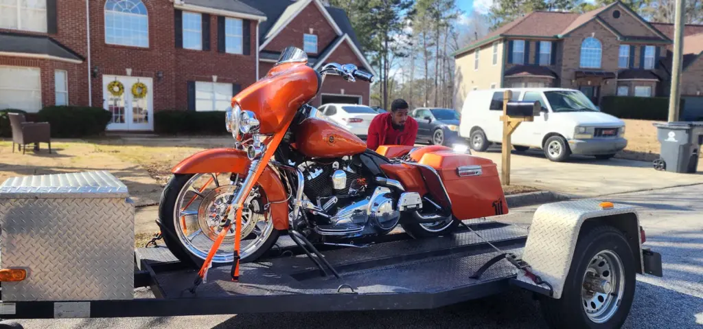 An orange motorcycle secured on a trailer sits in a suburban neighborhood with a person in red behind it.