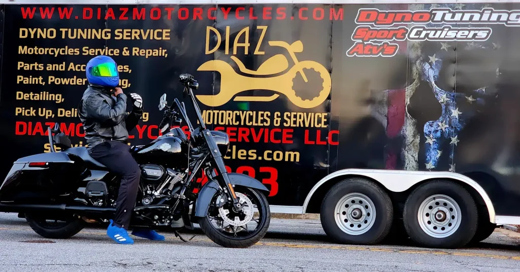 Rider in a blue helmet on a grey Harley bagger parked alongside the large Diaz Motorcycles enclosed trailer listing all the shop's services.