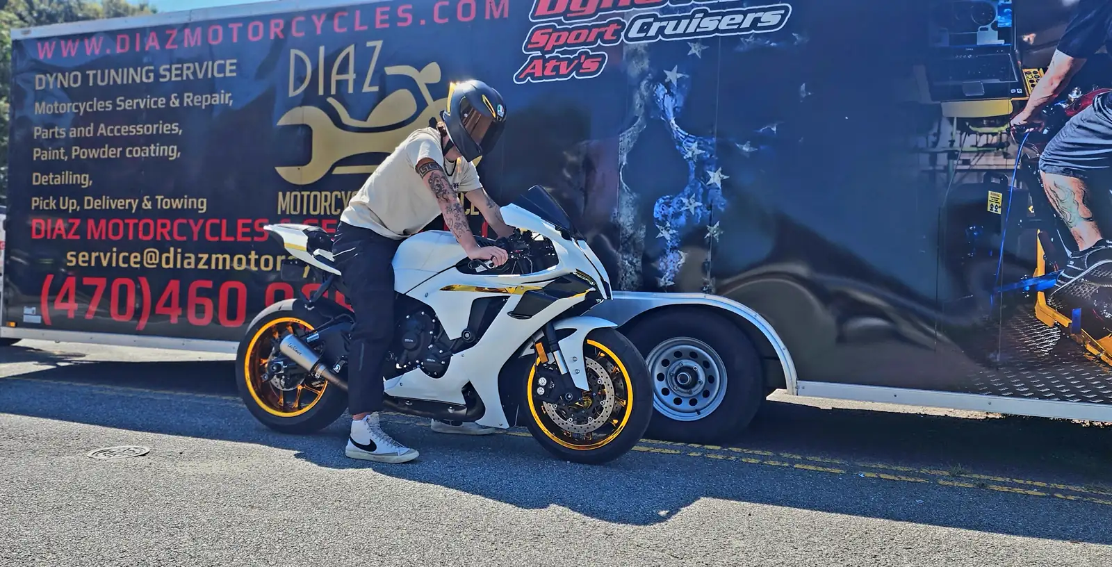Rider in helmet sits on a white and gold sportbike in front of the branded Diaz enclosed trailer parked at the curb.