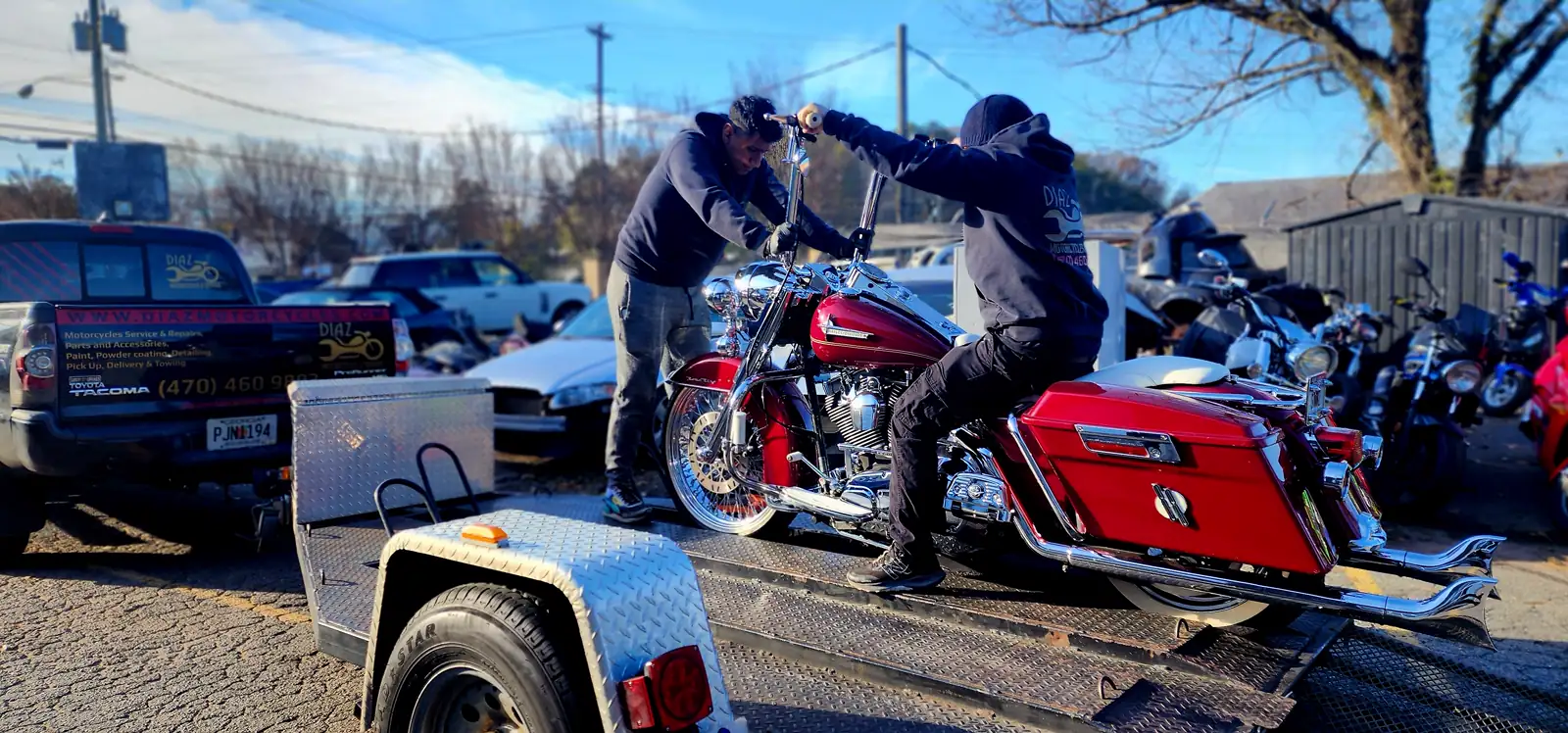 Two Diaz staff loading a red Harley-Davidson Road King onto an open trailer in bright sun, one steering from the saddle and one pushing from the front, branded Tacoma at left.