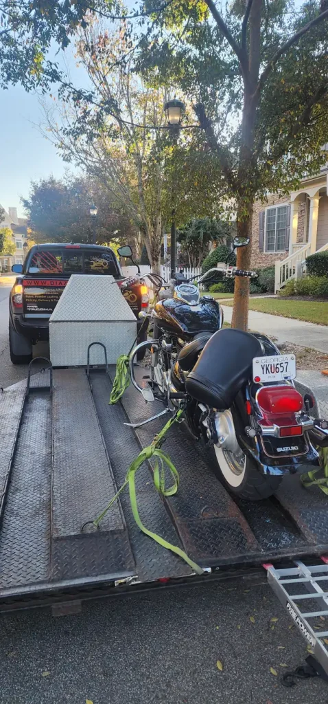 View from behind an open trailer loaded with a black Suzuki cruiser strapped with green straps, hitched to the Diaz-branded Tacoma on a residential street.
