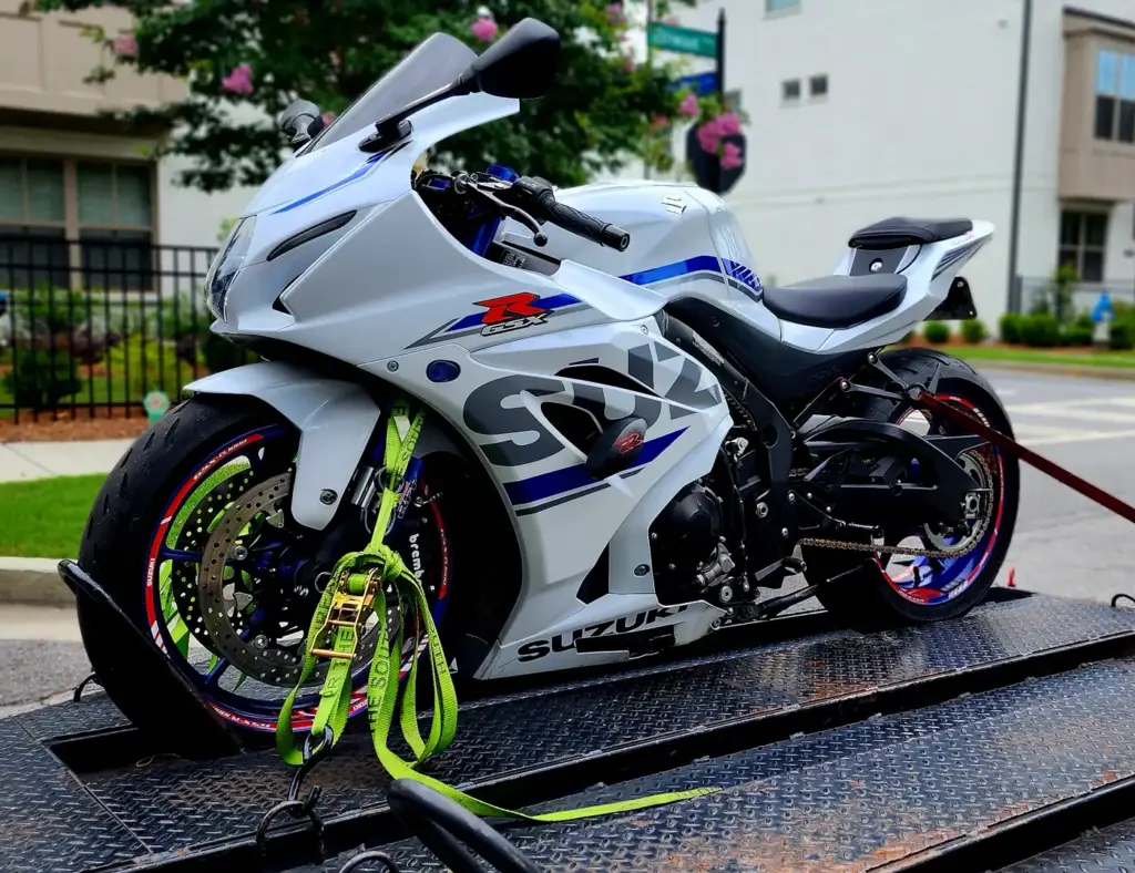 A white Suzuki GSX-R sports motorcycle is strapped with green ties on a flatbed trailer outdoors.