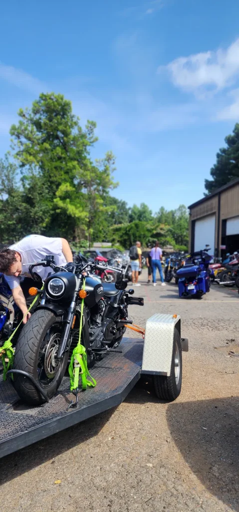 Worker in a white tee bends over a black Indian Scout Bobber strapped to an open trailer with green ratchet straps outside the shop.