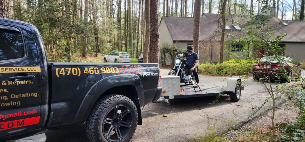 A man loads a motorcycle onto a trailer behind a black truck in a wooded residential driveway.